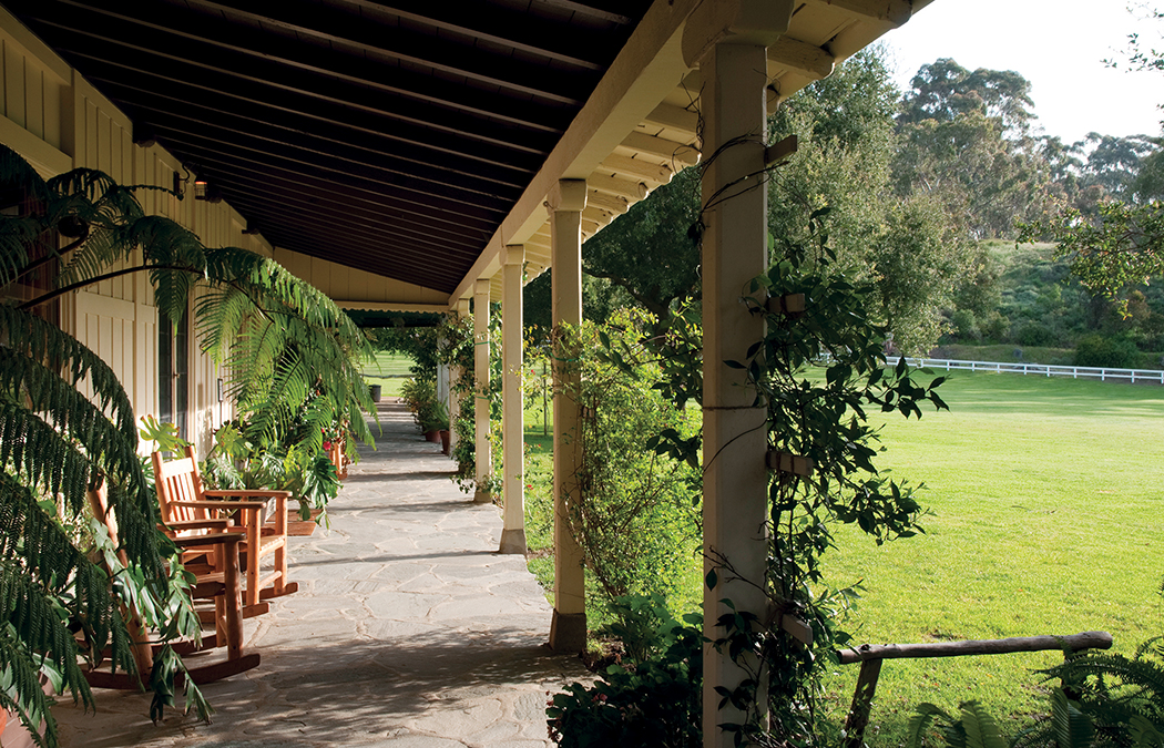 A peaceful porch with wooden rocking chairs, stone flooring, and lush greenery, opening to a sunny lawn and white fence.