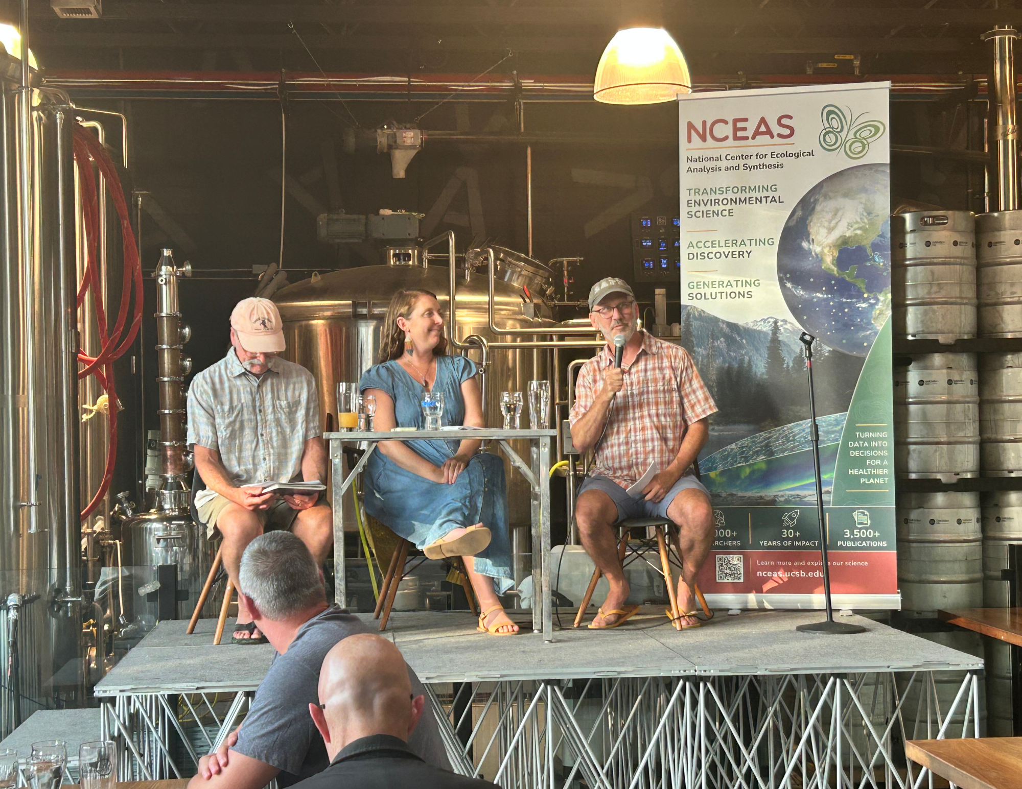 A panel discussion at a brewery features three speakers on stage, one using a microphone and two seated beside him. Brewing equipment is in the background, along with a banner promoting the National Center for Ecological Analysis and Synthesis.