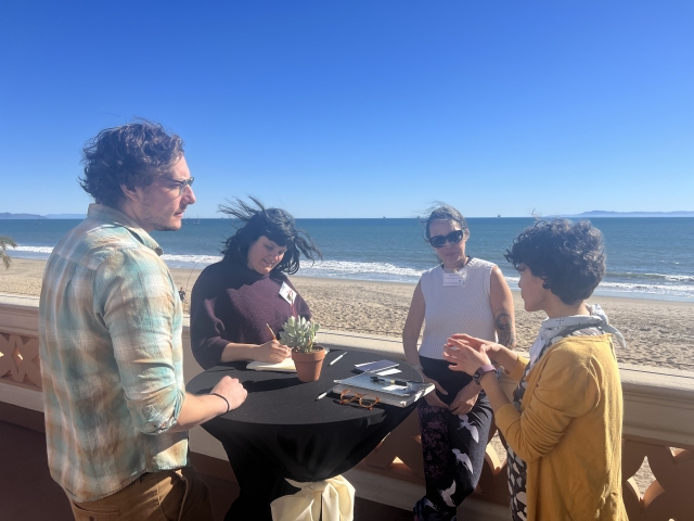Four people gather around a small circular table outside, with East Beach and the ocean behind them.