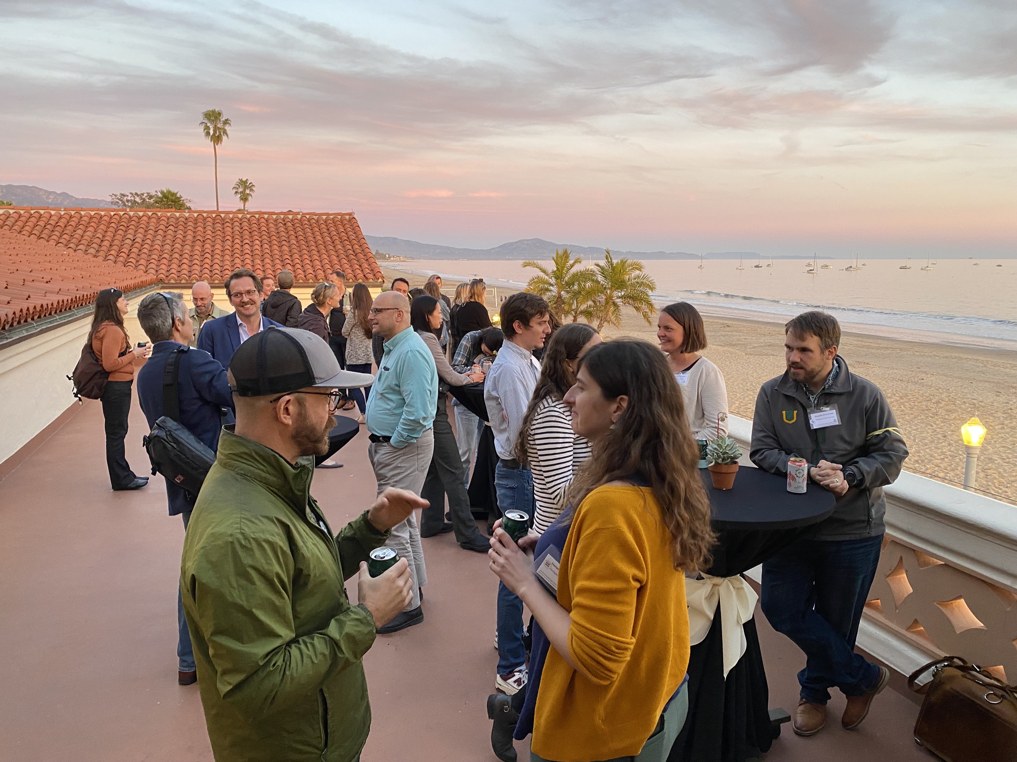 EDS Summit attendees gather for social hour on the patio of the Cabrillo Pavilion at sunset. In the background is East Beach, sailboats on the calm ocean, and a diminishing horizon line of mountainous coastline.