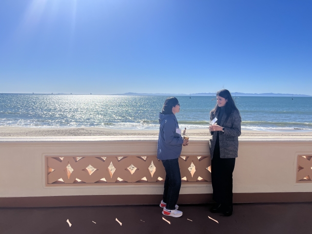Two Summit attendees talk together outside the Cabrillo Pavilion. In the background is the Pacific Ocean shining in the sunlight; the sky is cloudless.