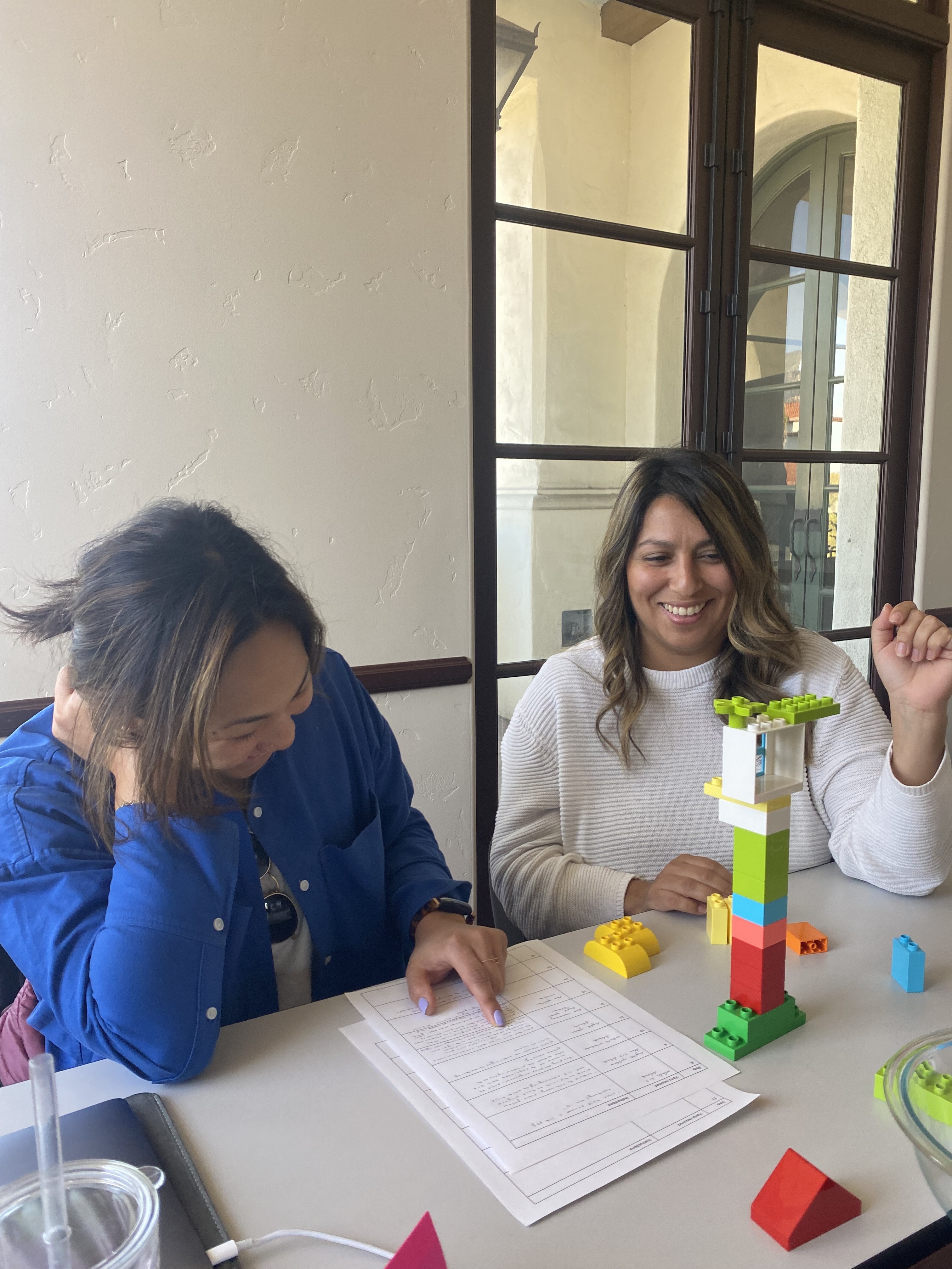 Two women sit at a table. One is assembling a structure made out of LEGOS blocks. The other is reading instructions from a piece of paper.