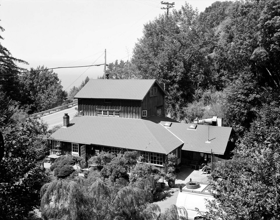 A two-story wooden house surrounded by trees, featuring large windows and a dirt driveway with a white vehicle parked nearby. The image is in black and white.