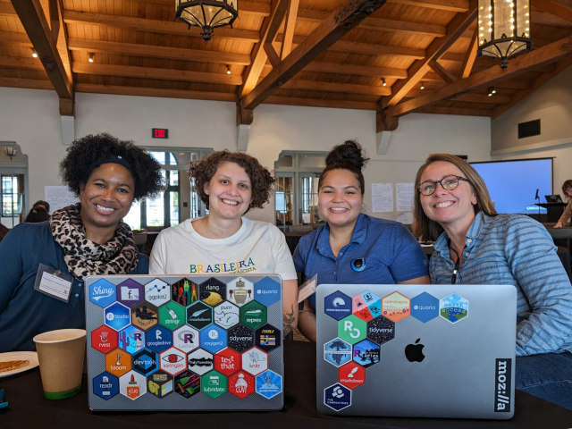 Four women sit together behind two open laptops displaying hex stickers. 
