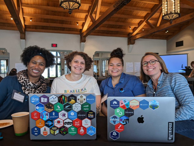 Four women sit together behind two open laptops displaying hex stickers.