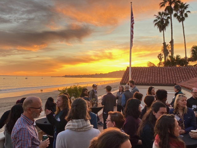 A group of EDS Summit attendees gather outside the venue at East Beach in Santa Barbara. In the background are a few palm trees and a vibrant sunset.