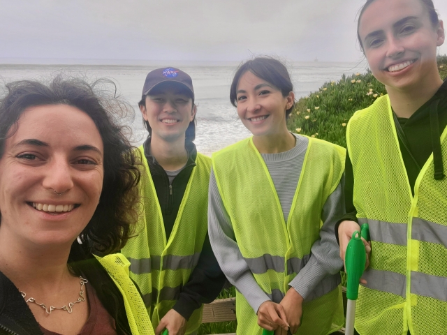 Four people in hi-viz vests stand near the ocean. 