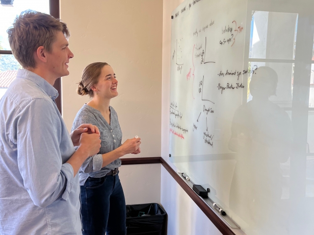 Two people stand in front of a whiteboard with writing on it. 