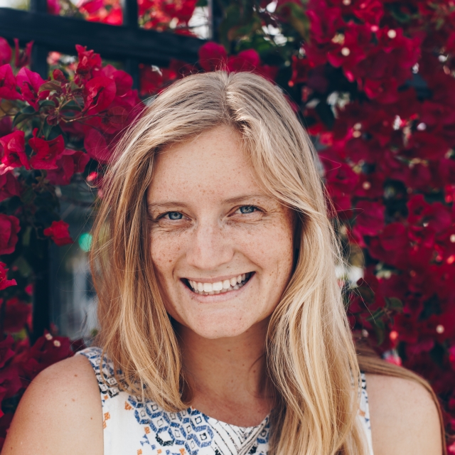 Alex, a fair skinned blonde-haired woman with blue eyes, is seen smiling in front of magenta flowers