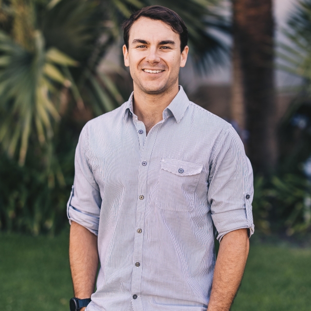 A headshot of Jake Eisaguirre in front of some dark green palm trees